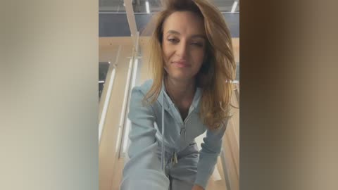 A video of a young woman with light skin and shoulder-length, wavy, light brown hair, smiling, wearing a light blue jumpsuit, standing in a modern, brightly lit room with large windows and wooden accents.