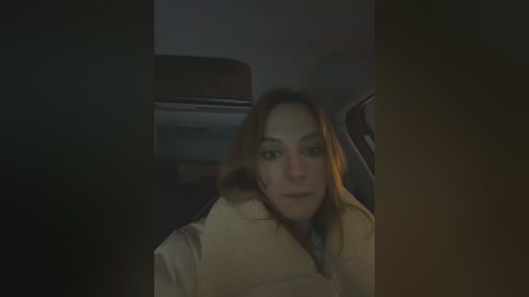 A dimly lit video of a woman with light brown hair, wearing a yellow bathrobe, sitting in a car. The background shows a dark interior with a visible part of a seat and a blurred air conditioning unit.