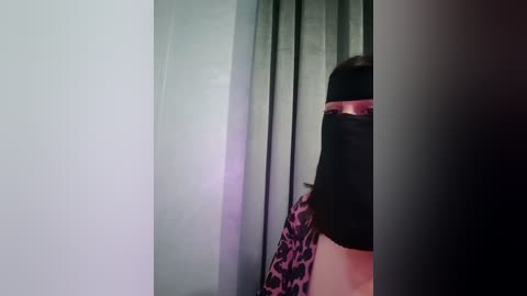 Video of a person wearing a black face mask, standing in a dimly lit room with a green curtain and leopard-print fabric in the background.