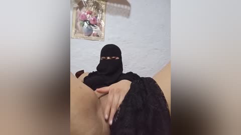 A video of a person with light skin, wearing a black hijab and a black lace dress, holding a hand over their crotch, in a dimly lit room with a white brick wall and a floral arrangement on the wall.