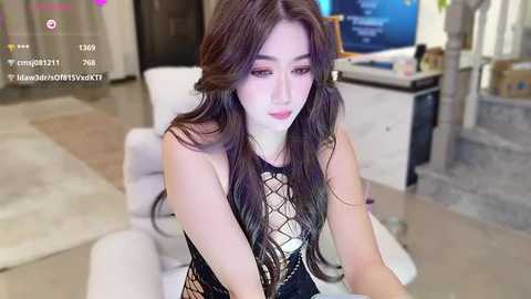 A video of an Asian woman with long, dark hair, wearing a revealing black lace bodysuit, sitting in a modern living room with a TV and white sofa.