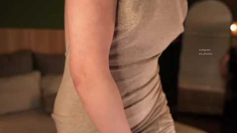 Video of a fair-skinned woman in a tight, glittery gold dress, posing in a dimly lit, cozy room with beige sofas and a wooden panel backdrop.