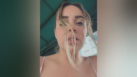 A close-up video of a young Caucasian woman with fair skin and blonde hair, wearing a pink spaghetti-strap top, against a teal and white background. Her face is partially obscured by her hair.