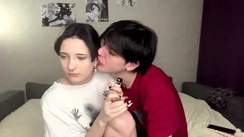 Media: Video of a young woman with long brown hair, wearing a white shirt, sitting on a bed. Another woman with short dark hair, wearing a red shirt, kisses her neck. Background features black-and-white photos on a beige wall.