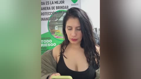 A video shows a young woman with long black hair, fair skin, and a black tank top, seated in front of a green and white poster.