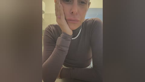 Video of a woman with light skin, dark hair, and a sad expression, wearing a long-sleeve, dark top and pearl necklace, leaning on her hand in a dimly lit bathroom.