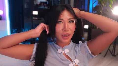 Video of an Asian woman with long black hair, wearing a blue cheongsam, posing in a modern, dimly lit room.