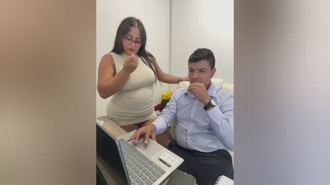 Video of a woman in a white tank top and glasses, and a man in a light blue shirt, both sitting at a desk, using a laptop.