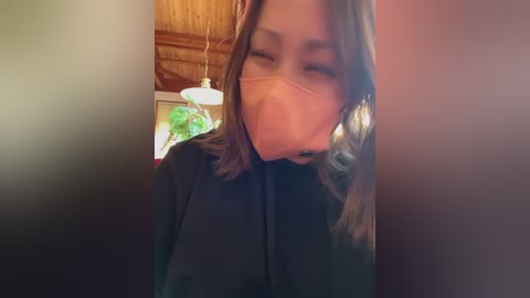 A blurry video of a woman with shoulder-length brown hair, wearing a dark robe, partially obscured by a blurred figure in the foreground. The background features a wooden ceiling and indistinct greenery.