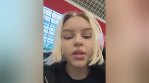 Video of a young woman with platinum blonde hair, light skin, and closed eyes, wearing a black top, in a modern indoor setting with a grid window and red wall.