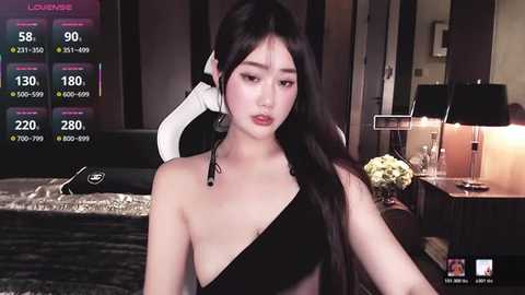 Video of an East Asian woman with long black hair, wearing a black one-shoulder dress, in a dimly-lit room with a bed, nightstand, and floral arrangement.