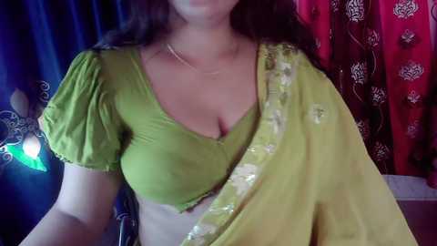 Video of a South Asian woman with medium skin tone wearing a green blouse and a yellow sari with gold embroidery. Background features blue and red curtains with intricate patterns.