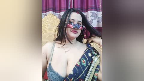 A video of a fair-skinned woman with long dark hair, wearing a colorful sari and a grey lace bra, holding a piece of fabric, against a maroon curtain backdrop.