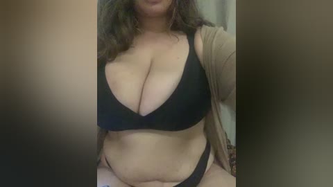 Video of a plus-sized woman with medium brown skin, wearing a black bra and panties, and a beige cardigan. Her long, wavy dark hair cascades around her shoulders. The background is blurry and neutral-toned.
