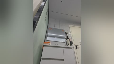 Video of a modern, minimalist kitchen with white cabinets, a stainless steel microwave, a black kettle, and a closed door. The room features soft, pastel green walls and a sleek, clean design.