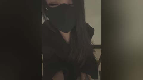 A dimly-lit video of a person wearing a black face mask and dark clothing, with long dark hair partially visible. The background is blurred and dark, suggesting a low-light indoor setting.