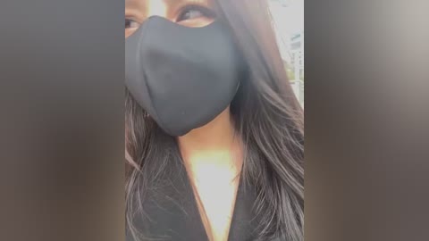 Video of a person with long, straight, dark brown hair, wearing a black face mask and black top. Background is blurred with a gradient from light to dark brown.