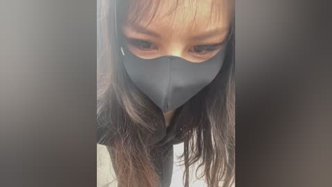 A close-up video of a woman with long, straight dark hair, wearing a grey face mask, and a black top. Her eyes are partially visible, and she gazes downwards. The background is blurred and dark.