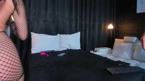 Video of a dimly lit bedroom with a black leather headboard, black bedspread, white pillows, a pink vibrator on the bed, and a woman in a fishnet top partially visible in the foreground.