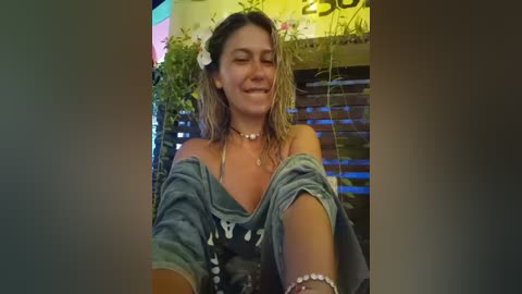 Video of a smiling, barefoot woman with wet, long, blonde hair adorned with a white flower. She wears a denim off-shoulder dress and beaded necklaces, seated in a lush, green outdoor setting with plants in the background.