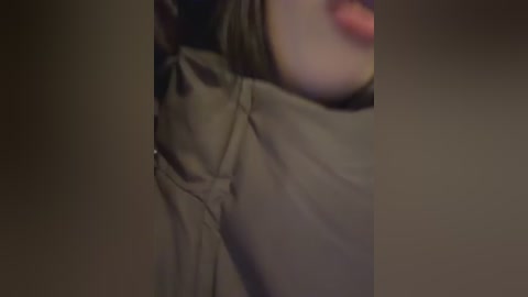 A dimly-lit close-up video of a woman's face and shoulder, partially obscured by a large, dark green blanket. Her lips are slightly parted, hinting at a casual or relaxed expression. The background is dark and indistinct.