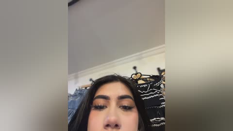 Video of a young woman with long dark hair, wearing black eyeliner and mascara, looking up. Background includes a white ceiling, a black and white patterned wall hanging, and a blue denim jacket.