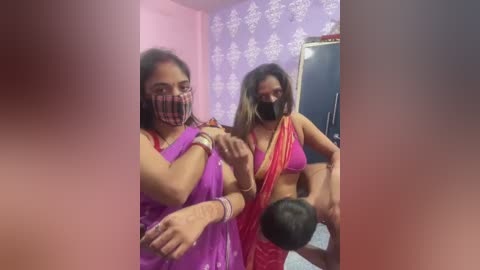Video of two women in traditional Indian attire, one wearing a purple sari and mask, the other in a red sari, posing playfully. Background features a purple floral wallpaper and a mirror.