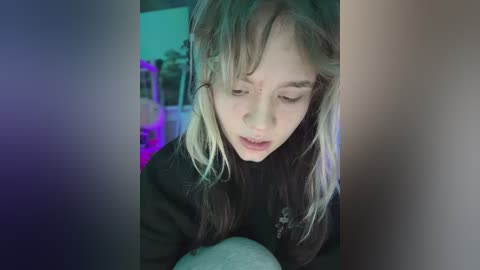 A young girl with messy, light brown hair, wearing a black sweater, sits on a white pillow, eyes closed, in a dimly lit room with purple and blue lighting.