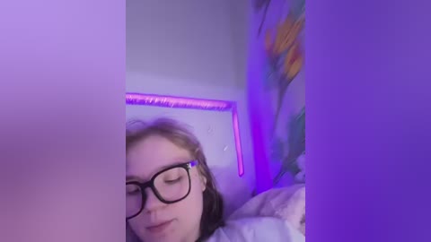 A video captures a young woman with fair skin, glasses, and light brown hair lying on a bed under purple lighting. Her face is partially obscured by a shadow, and she appears relaxed.