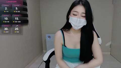 Video of an Asian woman with long black hair, wearing a teal tank top, white face mask, and seated on a white chair in a sterile room.
