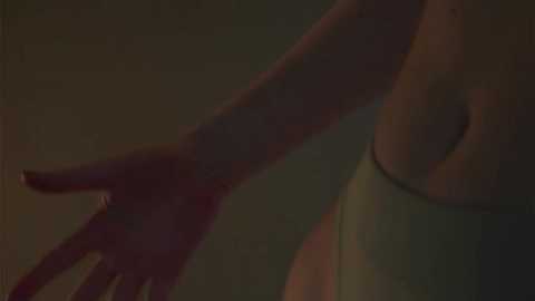 A dimly-lit video showing a close-up of a woman's arm, with her fingers slightly spread, against a blurred, dark background. She is wearing a white tank top, highlighting her slender, toned physique.