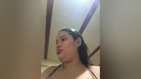 Video of a plus-size Latina woman with medium skin tone, long black hair, and full lips, wearing a black top, standing indoors with beige walls and wooden beams.