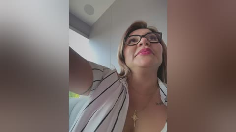 Video of a middle-aged woman with glasses, wearing a striped blouse, standing indoors near a window, looking upwards.