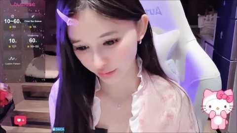 Video of an Asian woman with long black hair, wearing a pink blouse with frills, sitting in a white chair. Background includes a TV displaying streaming content and a pink Hello Kitty sticker.