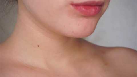 A close-up video of a person's neck and lower face, showing light skin with a mole, natural makeup, and slightly parted lips. The background is blurred and plain.