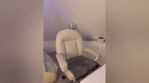 A video of a modern, minimalist bedroom with a white leather office chair, white bedside table, a bed with patterned duvet, and a glowing, starry wall.