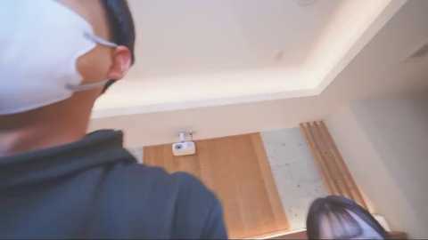 A video shows a close-up of a person in a white surgical mask, standing in a room with a wooden door, white ceiling, and a beige wall.