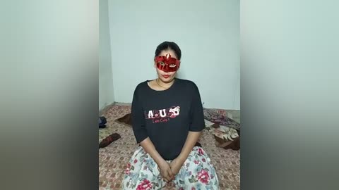 Video of a South Asian woman with medium-dark skin, wearing a black long-sleeve shirt with \"NOM NOM\" and a red floral mask, seated on a patterned bedspread, in a simple, dimly lit room.