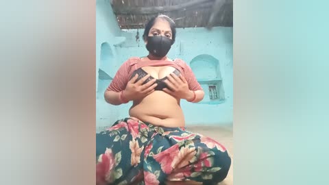 Video of a plus-sized woman with medium brown skin, wearing a black face mask, floral sarong, and a cropped red top, covering her black bra. She is seated in a rustic, light blue-walled hut with thatched roof.
