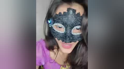 Video of a woman with long dark hair, wearing a black, intricate lace masquerade mask with silver sequins and a blue gem. She has a fair complexion and is dressed in a purple, off-the-shoulder top.