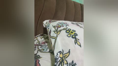 Video of a bed with a white pillow adorned with colorful floral patterns, and a matching duvet cover. The bed has a brown, tufted headboard. The background is blurred, with a hint of turquoise.