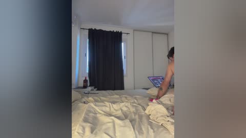 A video of a messy bedroom with beige sheets, a laptop on the bed, a person in the background, and dark curtains.