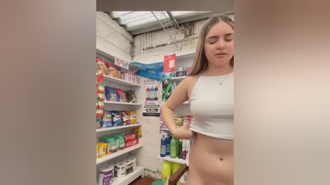 Video of a young Caucasian woman with light skin and long blonde hair, wearing a white crop top, standing in a cluttered, dimly lit convenience store with colorful shelves of snacks and beverages.
