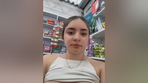 Video of a young woman with light skin, dark hair tied back, wearing a white tank top and silver necklace, standing in a brightly lit convenience store with colorful snacks and drinks on shelves in the background.