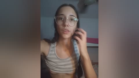 Video of a young woman with fair skin, long brown hair, and wearing glasses, holding her chin in a dimly lit room. She's in a gray and white striped tank top.