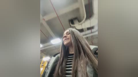 Video of a young woman with long brown hair and a striped shirt, smiling, standing in a dimly lit, industrial room with exposed pipes and yellow safety equipment.