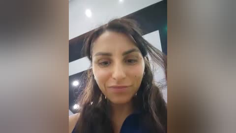 Video of a middle-aged woman with long, dark brown hair, wearing a blue shirt, looking slightly downward with a neutral expression. Background shows a modern, minimalist room with white and black walls and recessed ceiling lights.