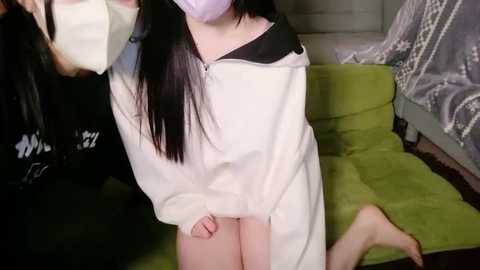 Video of an Asian woman in a white lab coat, mask, and long black hair, kneeling on a green couch in a dimly lit room with a bed and gray curtains.