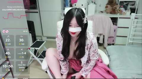 A video of an East Asian woman with long black hair, wearing a white face mask and a floral robe, sitting on a pink blanket in a cluttered bedroom.