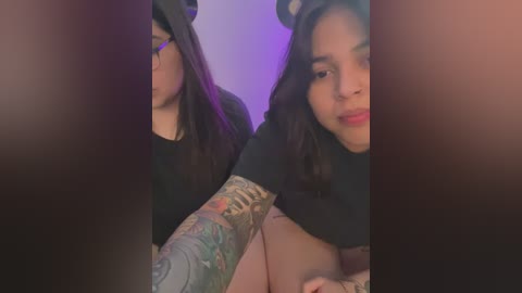 Video of a Latina woman with long, dark hair, wearing a black hat, glasses, and a black top. She has a colorful tattoo sleeve and sits in a dimly lit room with purple lighting.